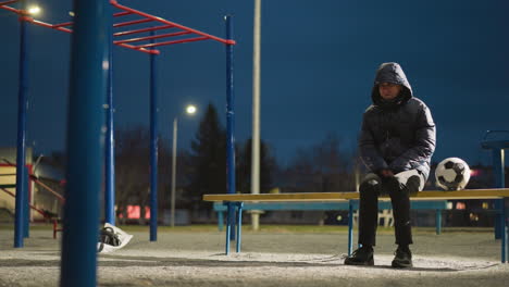 a man is seated outdoors at night in a quiet, empty stadium. he sits on a bench with his hands clasped between his legs, a soccer ball beside him