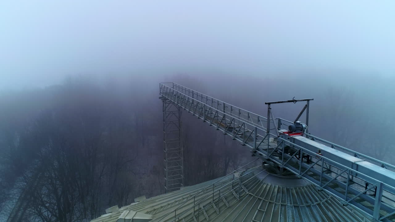 Sloping roof of an elevator tank with massive metal support over it. Drone footage along the metal pillar on the cistern. Foggy winter forest backdrop.