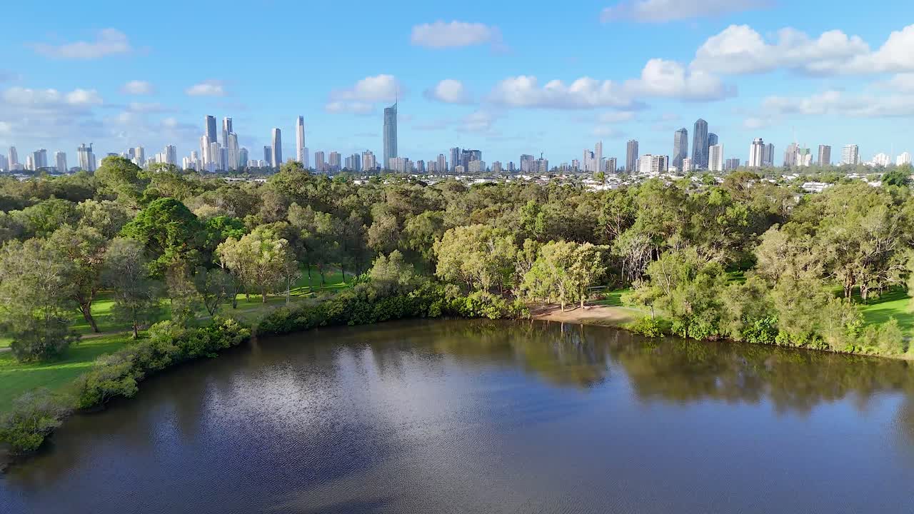 Drone footage captures lush parkland and city skyline under bright daylight in Gold Coast, Australia. Vibrant greenery and reflective water