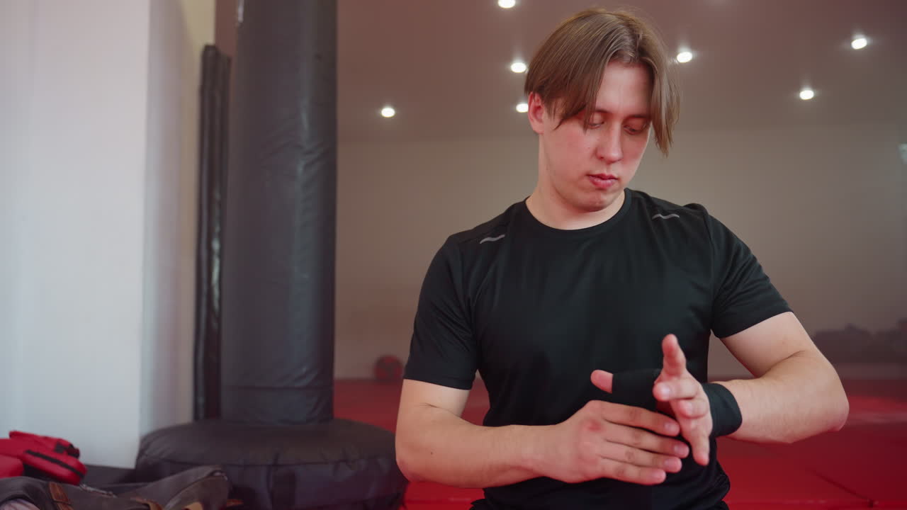 Boxer in black sportswear wraps his hand inside training gym, looking focused and alert before practice session, preparing mentally for sparring and combat