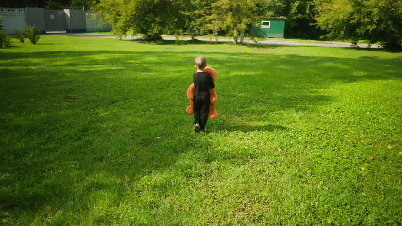 Rear view of small boy dressed in black carrying large orange teddy bear while walking across bright grassy field toward white security fence and green building surrounded by trees