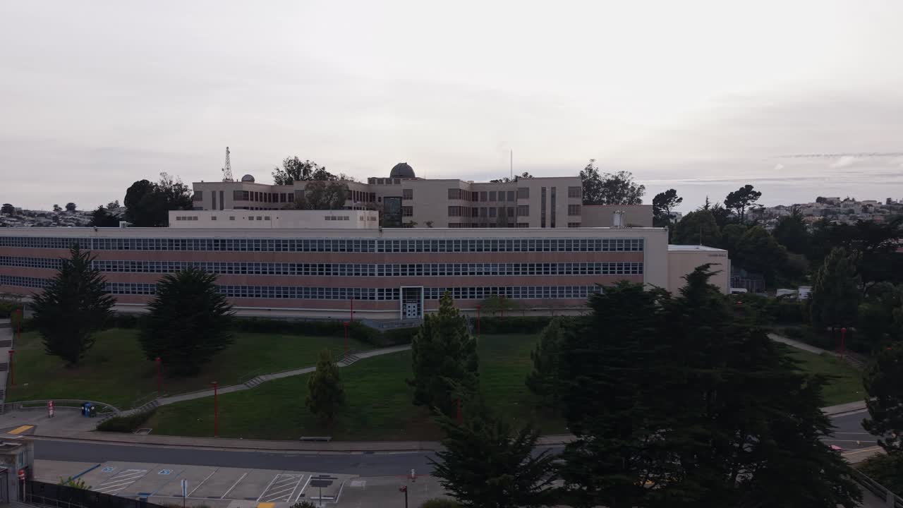 Vista aérea de seguimiento del campus principal del City College de San Francisco, mostrando su arquitectura moderna, la vegetación circundante y el estacionamiento bajo un cielo nublado con el paisaje urbano de fondo.