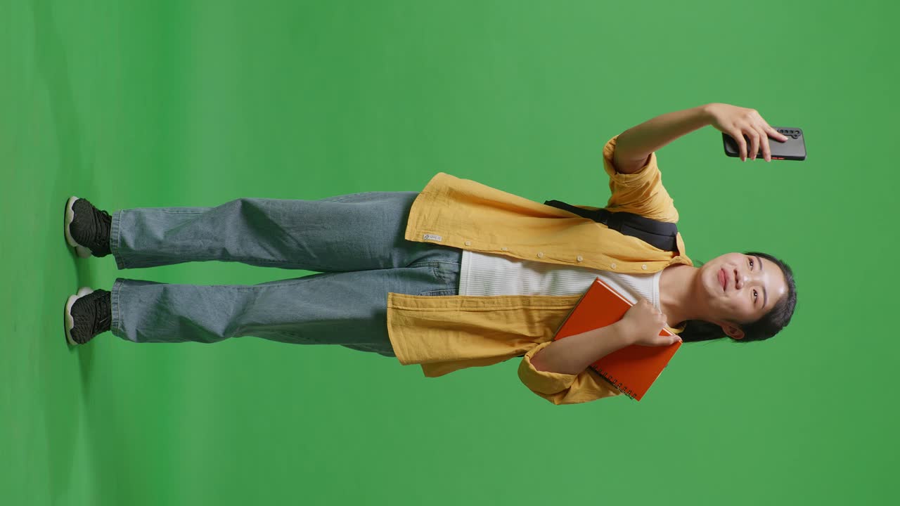 Full Body Of Asian Woman Student With A Backpack And Some Books Having A Video Call On Smartphone While Standing In The Green Screen Background Studio