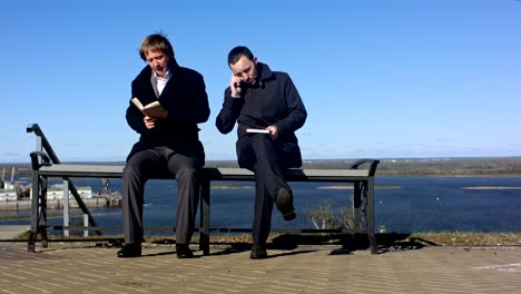 two men sitting outdoors on a bench with a book