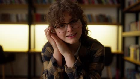 retrato de una feliz estudiante morena con gafas y una camisa a cuadros que se sienta en una mesa entre los libros de la biblioteca