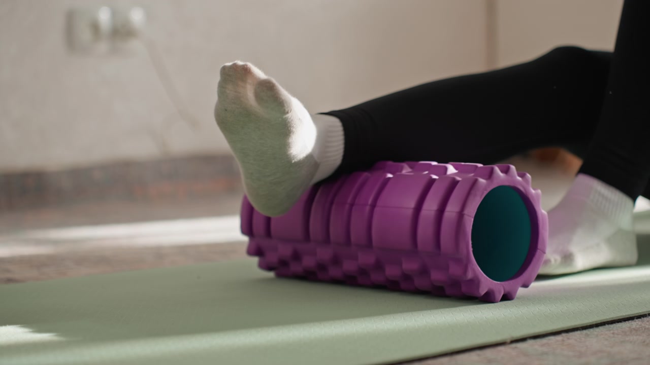 Home mat foam roller under calf during gentle physiotherapy session, black leggings and socks, purple textured roller on green mat, slow controlled movements for muscle release and improved mobility