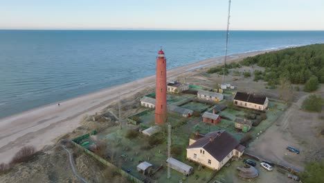 aerial establishing view of red colored akmenrags lighthouse, baltic sea coastline, latvia, white sand beach, calm sea, sunny day with clouds, wide drone shot moving forward