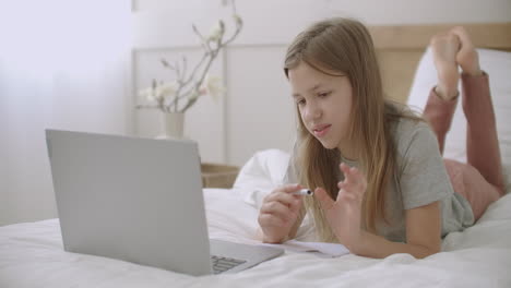 schoolgirl is communicating with teacher by video call on laptop lying on bed in home online lessons for secondary schoolers distant education