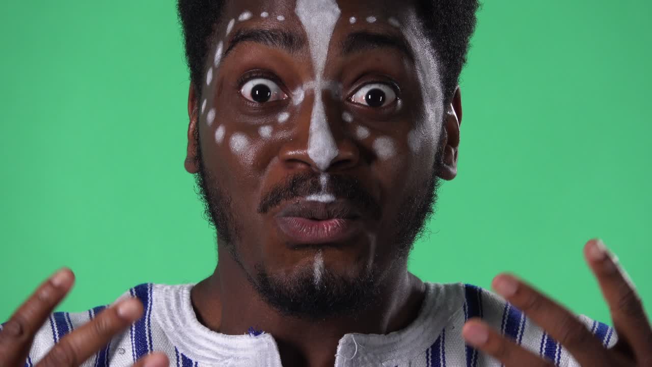 retrato de un joven afroamericano con una expresión facial sorprendida, luego muestra el pulgar como un signo. hombre negro con un patrón blanco étnico en la cara posando en pantalla verde en el estudio.