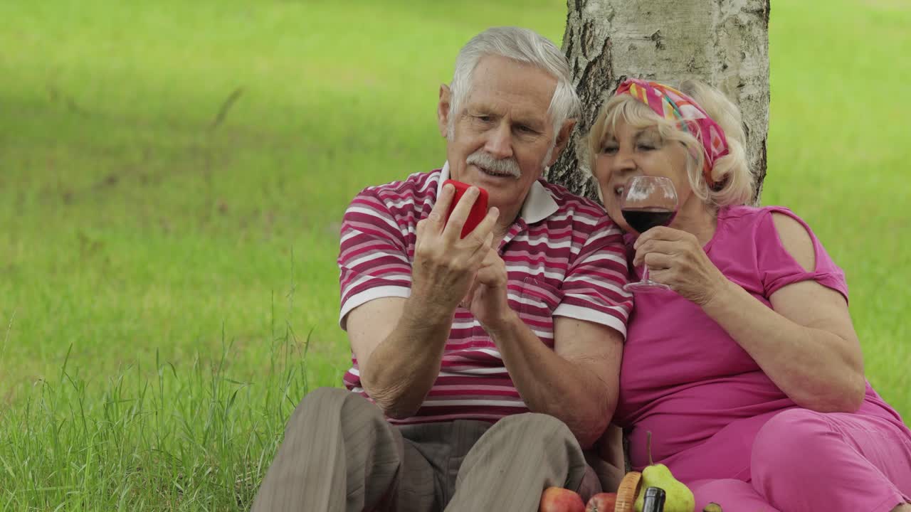 Family picnic. Senior old grandparents couple in park using smartphone online browsing, shopping