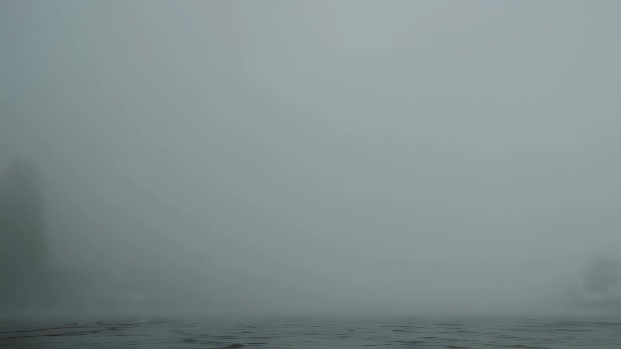 Dense fog drifting slowly across calm lake surface, gradually veiling distant shoreline trees and creating mysterious atmospheric landscape with soft, ethereal mood during quiet morning hours