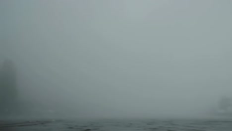 dense fog drifting slowly across calm lake surface, gradually veiling distant shoreline trees and creating mysterious atmospheric landscape with soft, ethereal mood during quiet morning hours