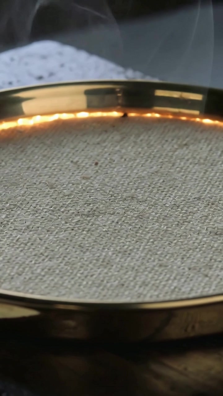 Video verticale: Braci ardenti su un bruciatore in ottone che producono fumo su un tavolo scuro, con un panno bianco sullo sfondo.