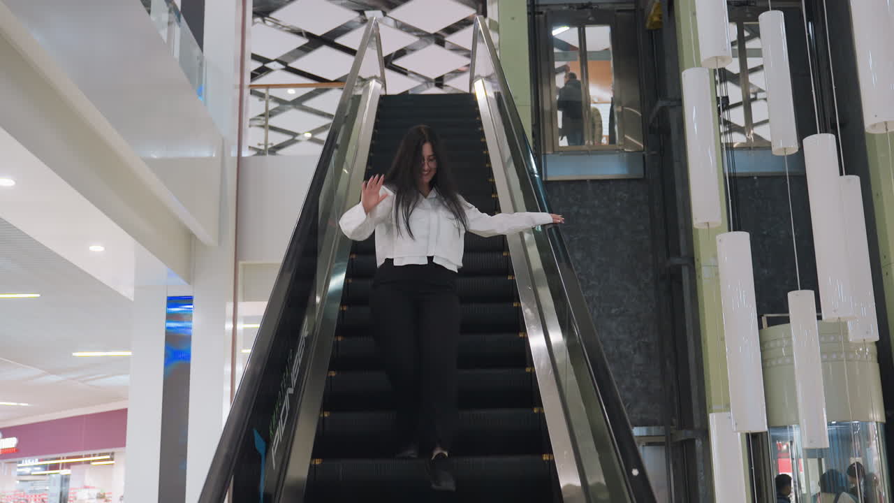 Young woman in white top and black pants descending escalator while smiling and waving toward someone in brightly lit modern mall interior with geometric ceiling and decorative hanging lights