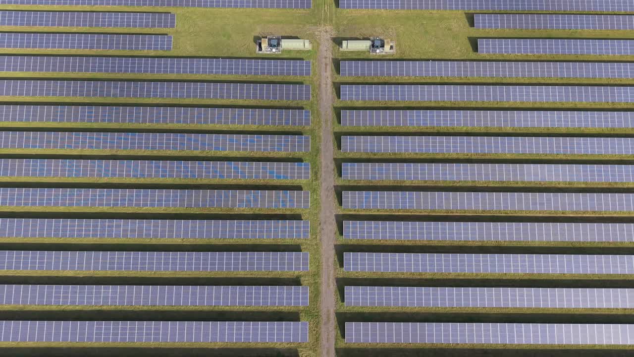 Aerial drone shot over a solar farm. Starts top-down, then reverses while tilting upward, revealing hundreds of panels, two electrical enclosures, and a cross-shaped gap between the rows of panels