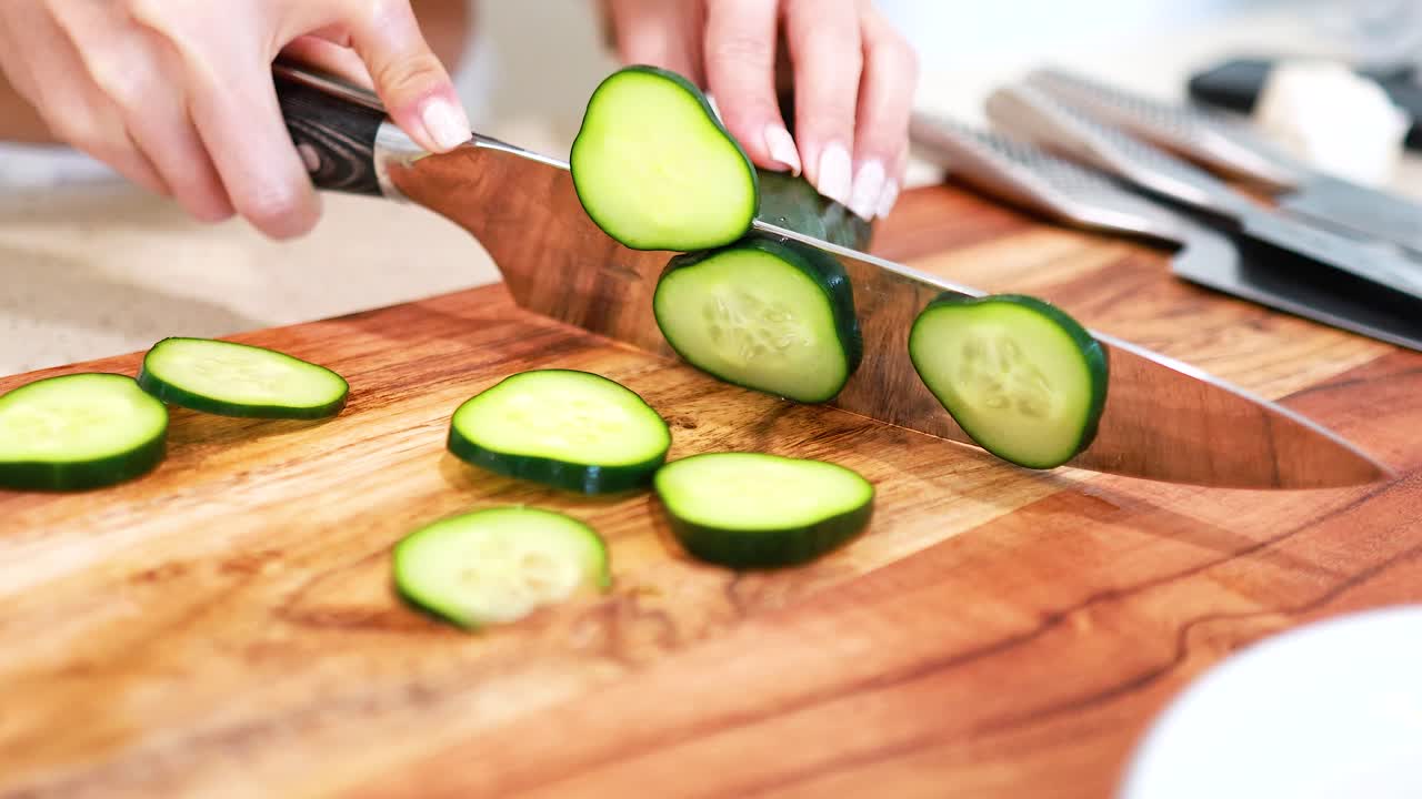 Hands slicing cucumber on a wooden board in a bright kitchen setting. Natural lighting and close-up composition enhance the focus on the action