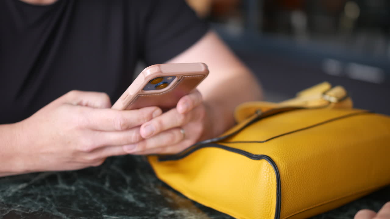 una mujer usando un teléfono inteligente rosa enviando mensajes de texto con ambas manos, sentada al lado de un bolso amarillo brillante en una mesa de mármol