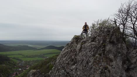 Aufschlussreicher-Reisender-Mit-Rucksack,-Der-Die-Spitze-Einer-Scharfen-Bergklippe-Erreicht,-Flache,-Farbige-Waldhügel,-Die-Ein-Kleines-Europäisches-Dorf-Im-Tal-Zeigen