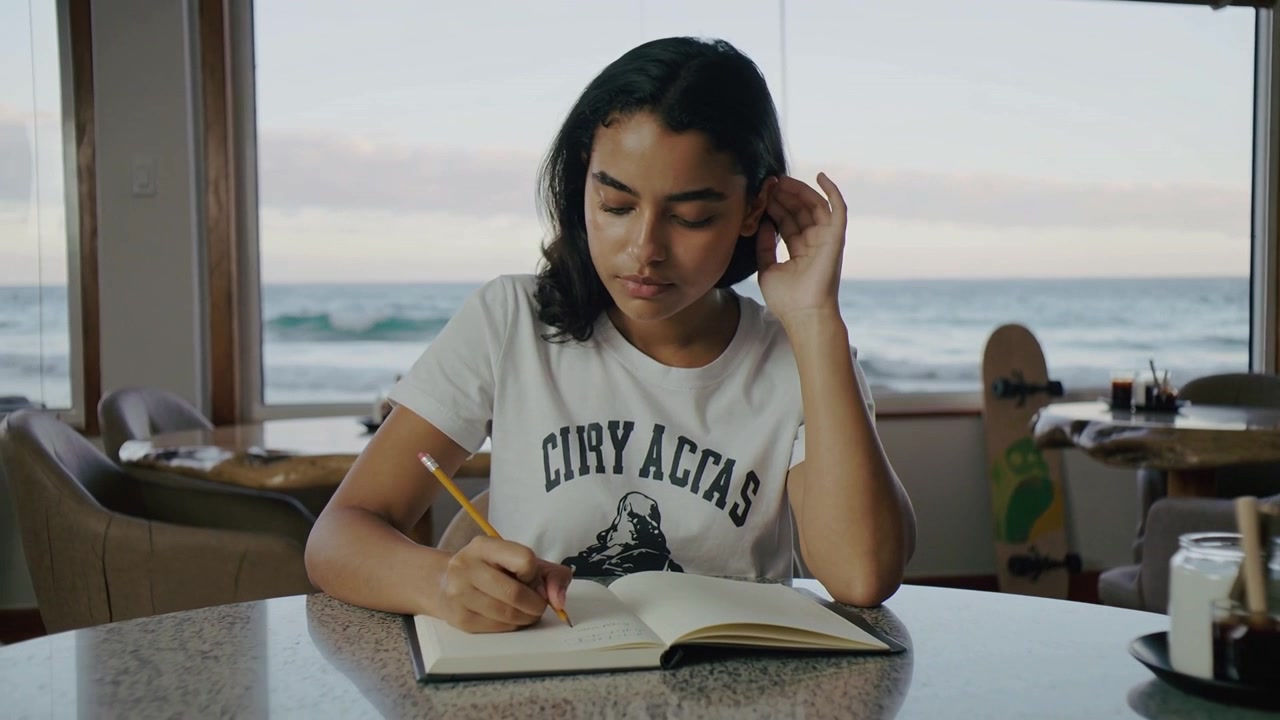 Donna che scrive, con una matita gialla pronta, prende appunti di studio in un caffè sulla spiaggia con un quaderno.