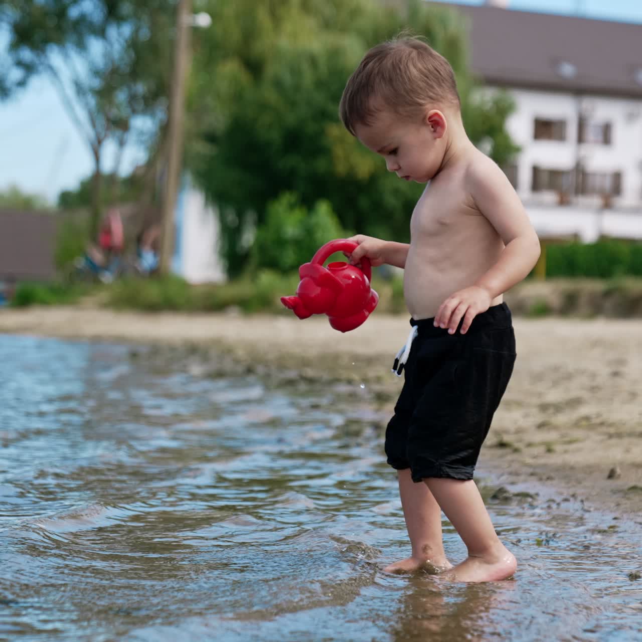 Caucasian toddler wearing black shorts filling the watering can. Baby boy playing near the river in summer