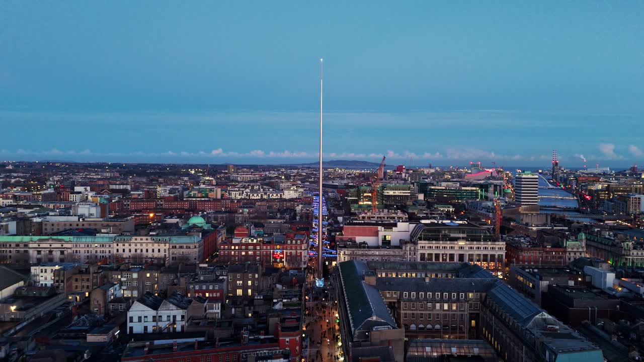 Luftaufnahme einer Landschaft mit einer Drohne, die sich von der Turmspitze in Dublin, Irland, bei Dämmerung in der blauen Stunde im Winter entfernt, mit irischen, beleuchteten Stadtgebäuden und belebten Straßen darunter mit TouristInnen