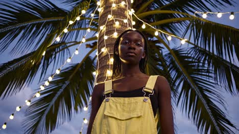 young woman in yellow overalls under palm tree at night