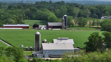 picturesque american farm with silos and barns, set amidst lush green fields. surrounded by vibrant trees and rolling hills, it embodies rural charm and agricultural lifestyle. aerial approaching.