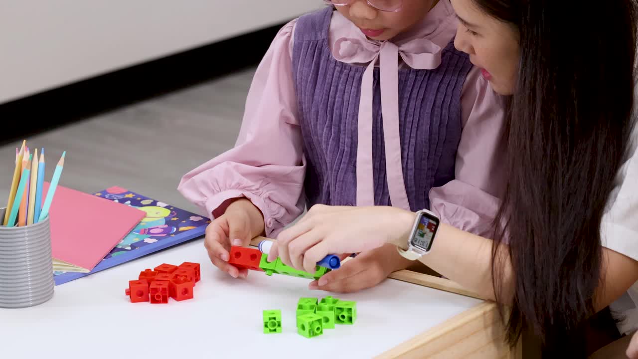 Adult guides young student stacking colored counting cubes on classroom table, static close-up shot