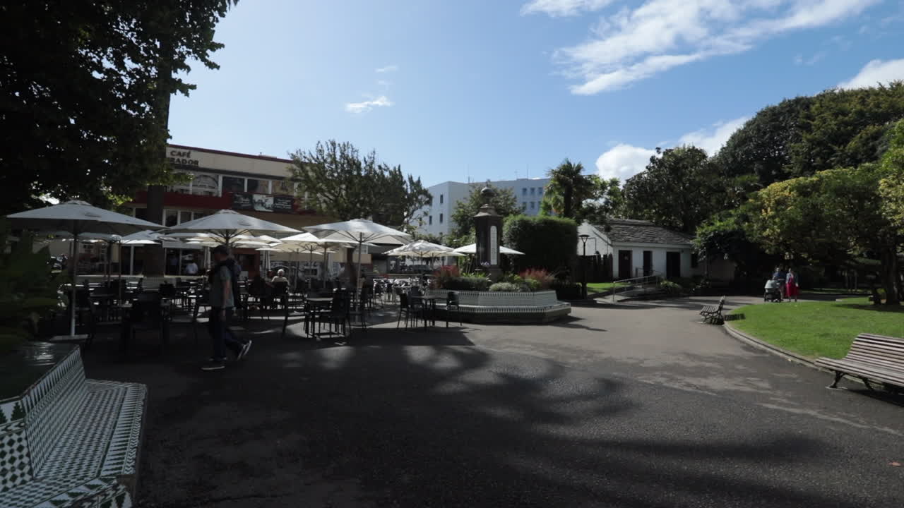 People walk in a sunny outdoor café courtyard surrounded by greenery and trees