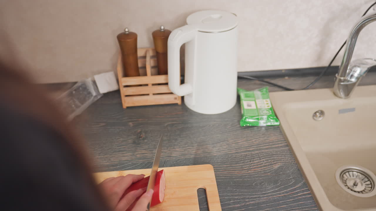 rear view of woman removing hotdog from plastic wrapper placing it on wooden cutting board preparing to slice with kitchen knife during meal prep in modern kitchen beside kettle