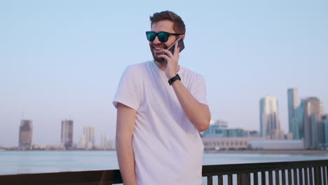 happy handsome man in sunglasses and white t-shirt with bristles talking on the phone standing on the waterfront in the summer against the city and buildings