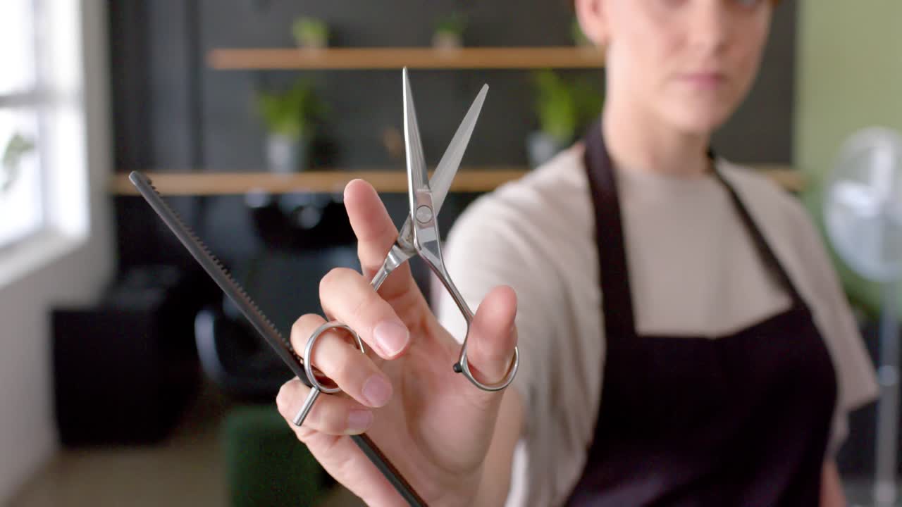 Midsection of caucasian female hairdresser in black apron holding scissors and comb, in slow motion