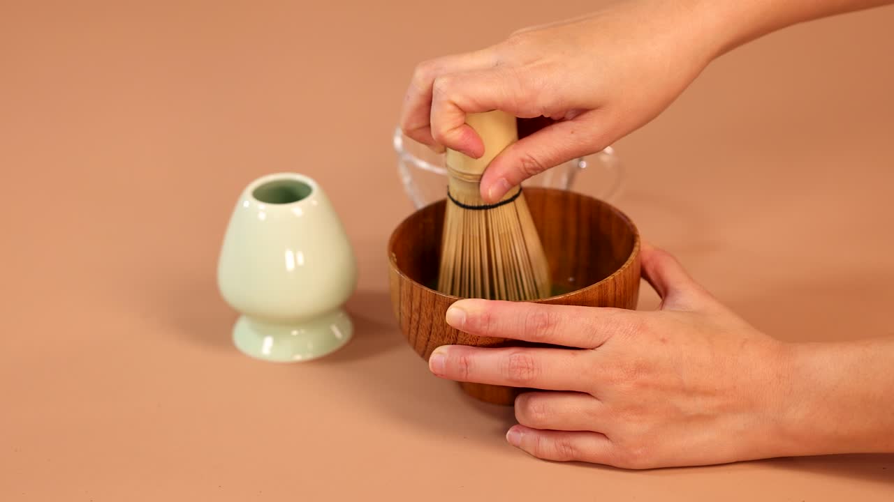 Hands whisk matcha green tea in wooden bowl using traditional bamboo whisk, minimal studio lighting