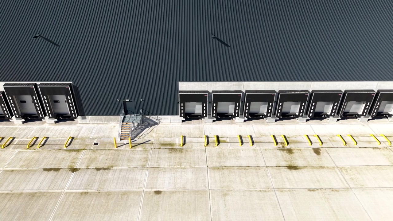 Dynamisch laagvliegend luchtbeeld langs de industriële betonnen platforms en roldeuren van een magazijn distributiecentrum