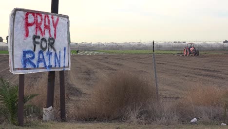 Ein-Traktor-Pflügt-Während-Einer-Dürrezeit-In-Kalifornien-Ein-Sehr-Trockenes-Feld,-Während-Ein-Schild-Die-Menschen-Auffordert,-Für-Regen-Zu-Beten