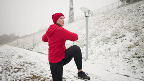 coach works out outdoors, lifting her leg on snowy ground near stairway, wearing red hoodie and black gloves, surrounded by snowy hill, pathway, lamppost, and foggy atmosphere