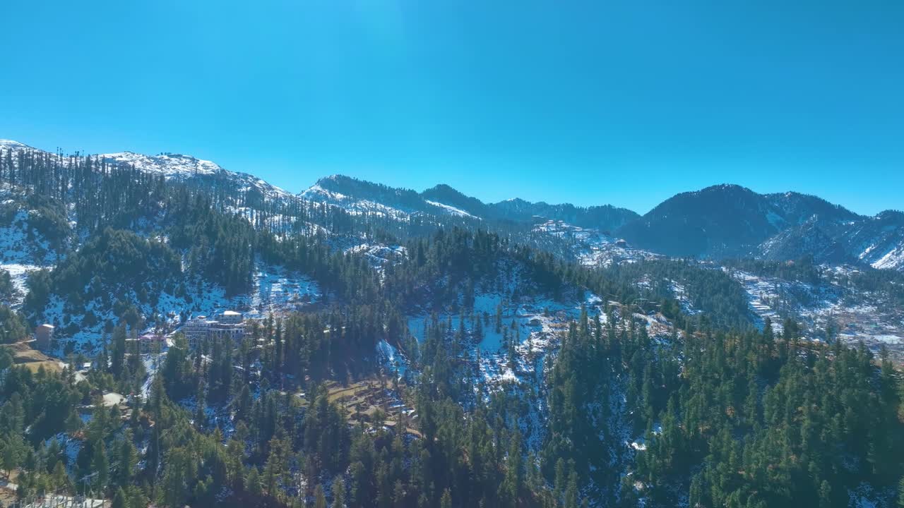 An aerial view of a snowy mountain range, with scattered buildings nestled among the forested slopes
