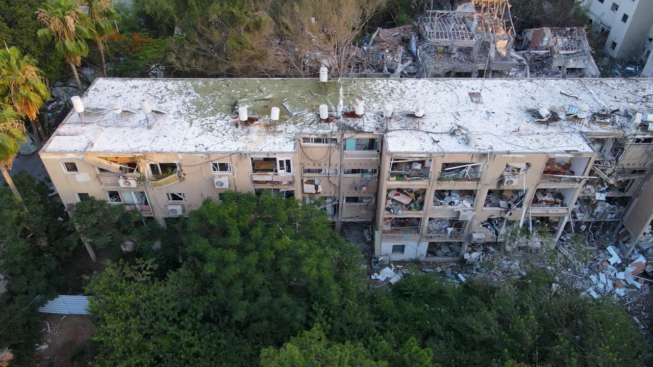 Top-down drone view of a destroyed building in Tel Aviv. Blown-out facade, damaged interior, debris on the ground, and another ruined building in the background