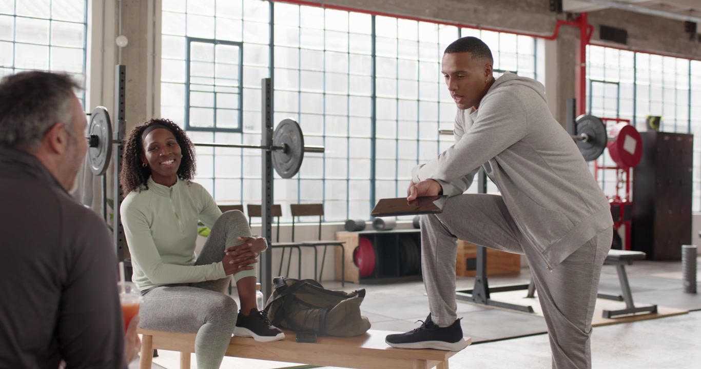 Male trainer in gray tracksuit stepping on bench, tapping tablet and explaining workout plan in gym