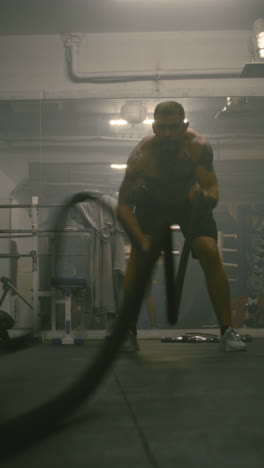 professional male boxer does cardio or endurance workout before championship fight. athletic man exercises with battle ropes in dark boxing gym. vertical shot