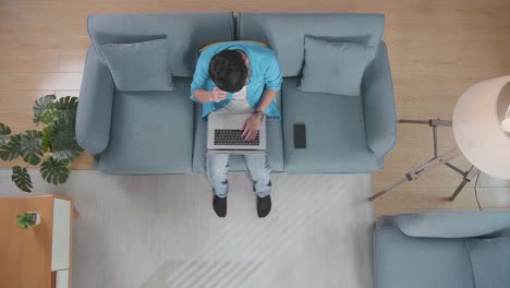 top view zoom out shot of a tired man having a headache while sitting on the sofa with a smartphone typing on a laptop at home