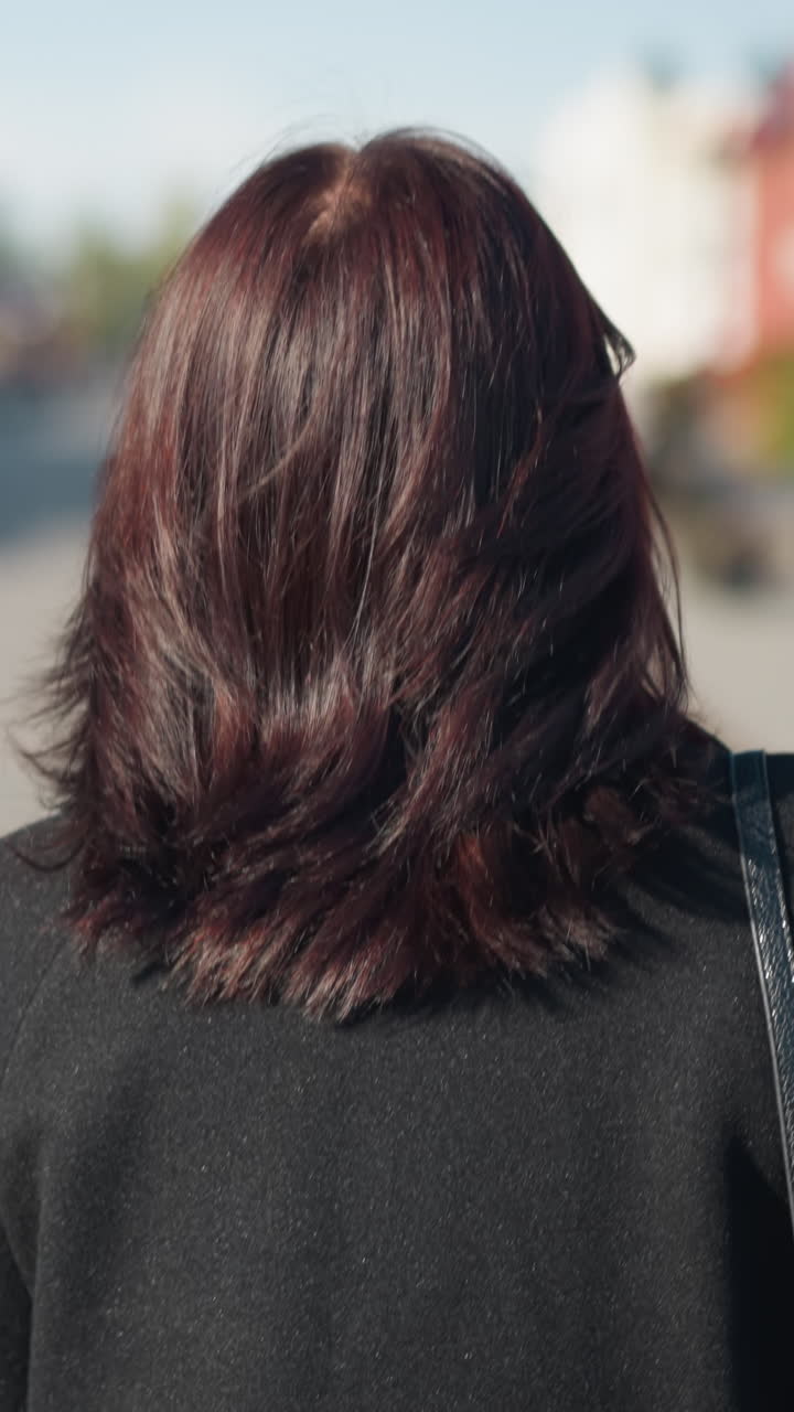 흑색 코트를 입고 다니는 갈색 머리카락의 여성의 근접 후면 모습, 이 많은 도시 거리에서 촬영되어 일상적인 우아함을 보여줍니다.