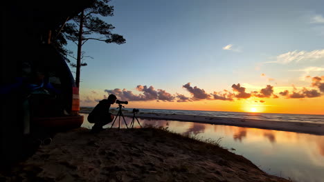 silhouette of a person holding camera on tripod stand at sunset in public camping at irbe mouth in latvia. static shot
