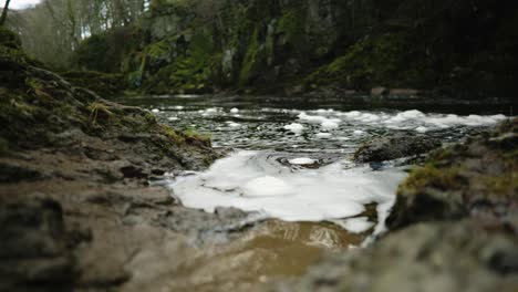 Schaumblasen,-Die-Auf-Der-Oberfläche-Des-Dunklen-Wassers-Im-North-Esk-River-In-Schottland-Schwimmen,-Schwappen-Langsam-Gegen-Das-Flussufer,-Während-Ein-Schnell-Fließender-Fluss-An-Einer-Moosbedeckten-Klippe-Im-Hintergrund-Vorbeirauscht