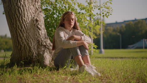 woman sits on grassy ground with eyes closed, resting against tree trunk, sunlight reflecting off her peaceful face, her arms are folded over her knees, with blurred background poles and stadium
