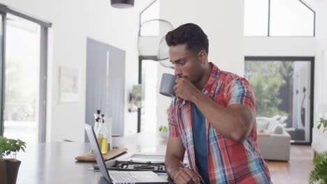 Biracial-Mann-Trinkt-Kaffee-Und-Benutzt-Laptop-In-Sonniger,-Moderner-Wohnung,-Zeitlupe