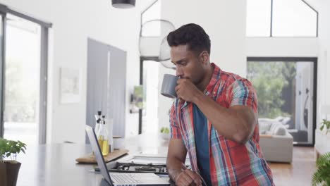 hombre biracial tomando café y usando computadora portátil en un apartamento moderno soleado, cámara lenta