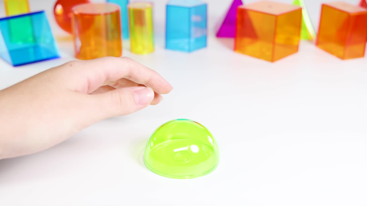 A hand manipulates vibrant 3D geometric shapes on a white surface, highlighting educational play and tactile exploration