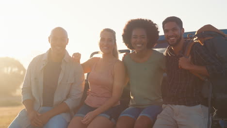 Portrait-Of-Friends-Sitting-On-Tailgate-Of-Pick-Up-Truck-On-Road-Trip-To-Cabin-In-Countryside