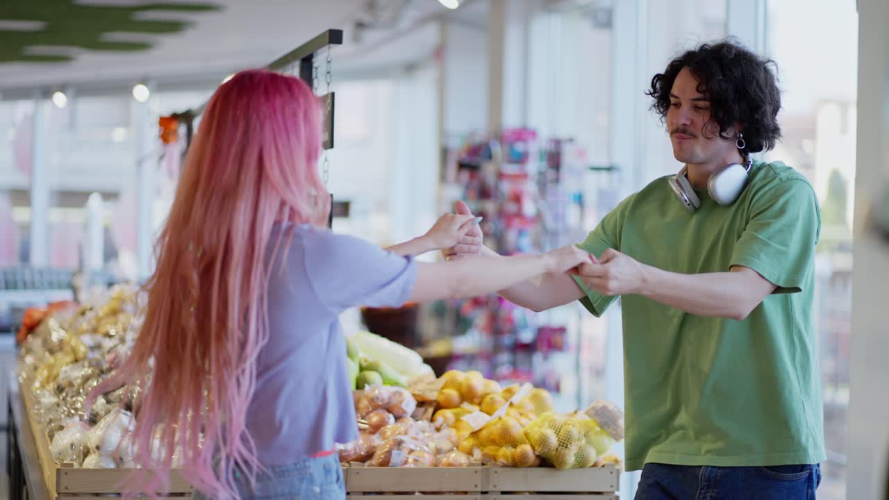 A happy girl with pink hair dances with a brunette guy in a green T-shirt near the counter in a supermarket